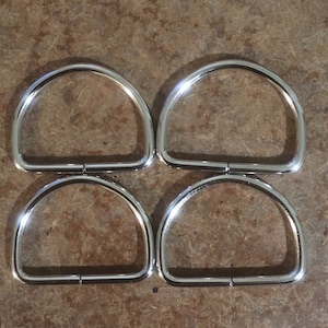 May include: Four silver D-rings, each with a smooth, rounded surface. The rings are arranged in two rows of two, with the top row slightly offset from the bottom row.