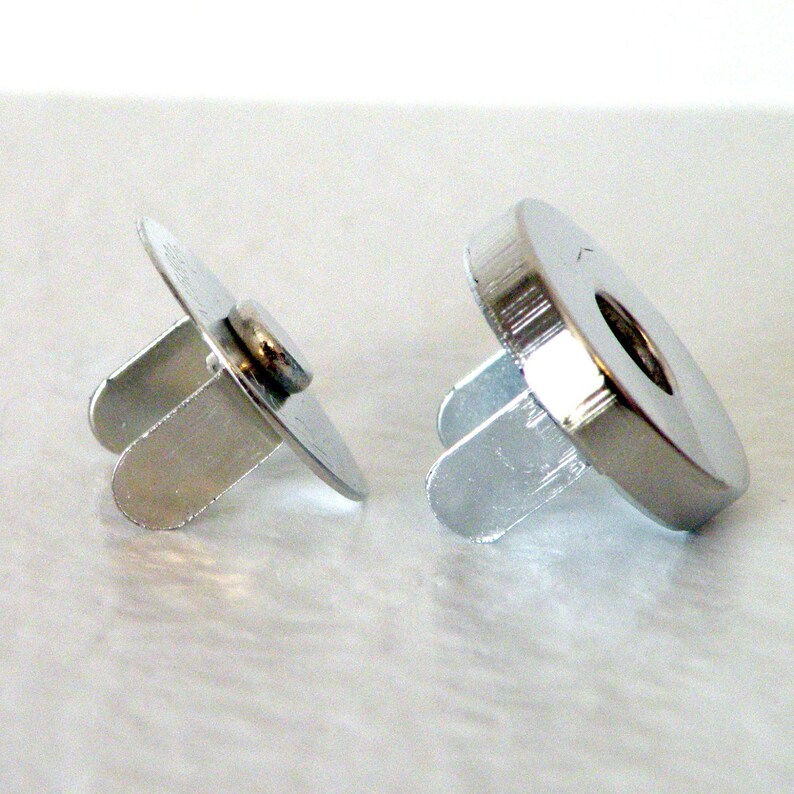 May include: Two silver magnetic closures for clothing. The closures have a round base with a small hole in the center and two prongs extending out from the base.