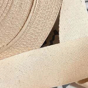 May include: Close-up of a roll of natural cotton webbing. The webbing is woven with a tight, even weave and has a slightly rough texture.