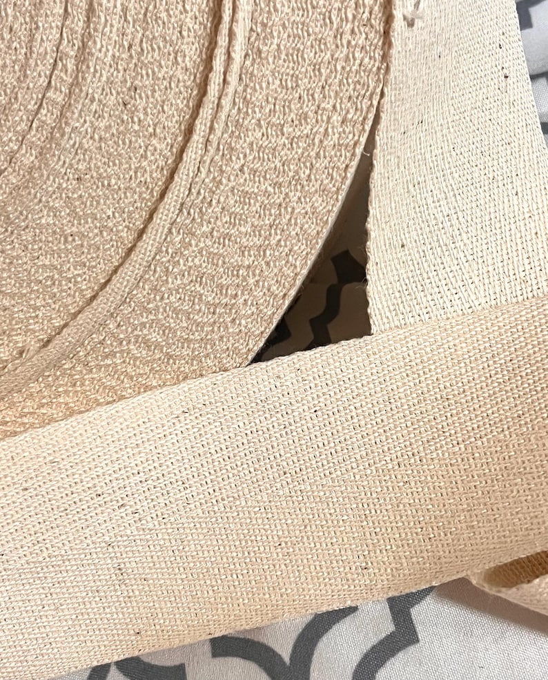 May include: Close-up of a roll of natural cotton webbing. The webbing is woven with a tight, even weave and has a slightly rough texture.