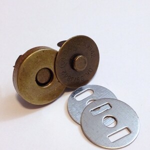 May include: A set of three magnetic closures for sewing projects. The closures are made of metal and have a brass finish. The larger closure has a round shape with a raised center and the text "OKP TAI 519842" printed on the side. The smaller closures are round with a hole in the center and a rectangular cutout on the side.