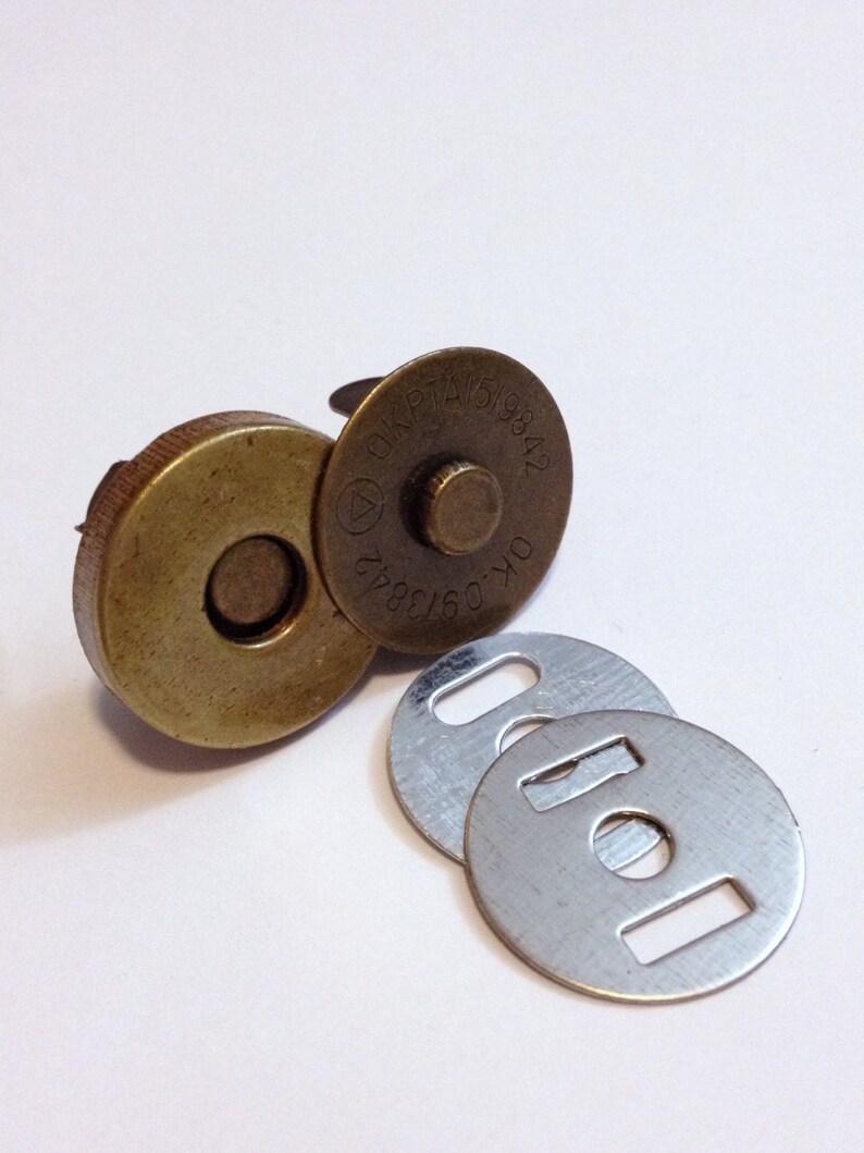 May include: A set of three magnetic closures for sewing projects. The closures are made of metal and have a brass finish. The larger closure has a round shape with a raised center and the text "OKP TAI 519842" printed on the side. The smaller closures are round with a hole in the center and a rectangular cutout on the side.