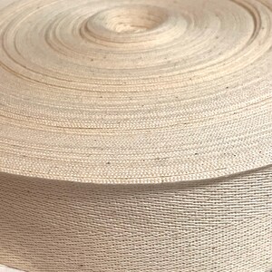 May include: A close-up of a roll of natural cotton twill tape. The tape is tightly wound and has a smooth, even texture.