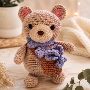 May include: A crocheted teddy bear in light brown yarn with pink inner ears and black button eyes. The bear wears a light purple crocheted scarf and carries a small brown crocheted bag containing light purple crocheted flowers across its body.
