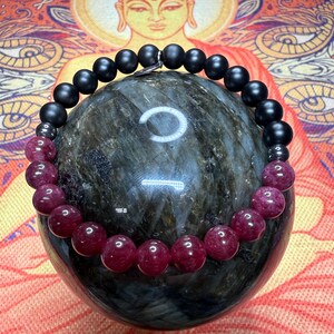 May include: A beaded bracelet featuring a sequence of matte black spherical beads and polished deep red translucent beads. The bracelet includes small metallic spacer beads and a small silver-toned charm, arranged in a circular design.