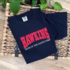 May include: A navy blue short-sleeve t-shirt featuring the red text HAWKINS in a collegiate font, with the white text HOME OF THE UPSIDE DOWN printed below it. The shirt has a Comfort Colors brand label at the inner neckline showing size L.
