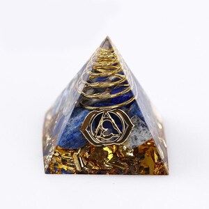 May include: A pyramid-shaped object made of clear resin containing blue lapis lazuli stones, gold-coloured metallic flakes at the base, a coiled gold-coloured wire spiral, and a gold-coloured metal emblem featuring a triangular symbol on the front.