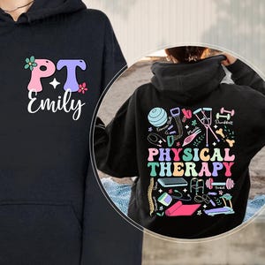 May include: A black hooded sweatshirt featuring a front graphic with the text PT Emily in pink and purple letters with small flowers. The back design displays the text PHYSICAL THERAPY in colorful letters surrounded by various exercise equipment icons.