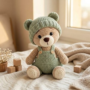 May include: A crocheted teddy bear with light brown yarn, wearing a matching sage green beanie with bear ears and sage green overalls featuring two small wooden buttons. The bear has a cream-colored muzzle, a dark brown nose, and black eyes.
