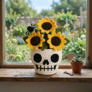 May include: A crocheted off-white skull planter containing three yellow sunflowers with brown centers and green leaves. A small black spider is attached to one sunflower. Next to the skull is a small terracotta pot with a succulent and a metal key.