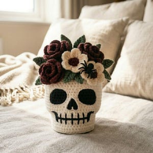May include: A crocheted skull planter in off-white yarn with black stitched eye sockets and a mouth. The top is filled with a bouquet of dark burgundy and cream crocheted flowers, green leaves, and a small black spider accent attached to a petal.