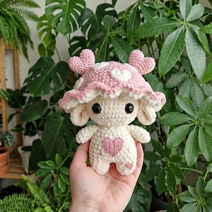 Puede incluir: Un peluche de ganchillo con cuerpo color crema, un corazón rosa en el pecho y un sombrero en forma de seta rosa adornado con corazones blancos. La figura tiene grandes ojos negros, orejas pequeñas y dos protuberancias en forma de corazón.