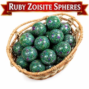 Op de afbeelding: Een gevlochten rieten mand gevuld met meerdere gepolijste robijn-zoisiet bollen. Elke bol heeft een gevlekte groene basis met duidelijke magenta en paarse insluitingen. De tekst "RUBY ZOISITE SPHERES" staat in het wit op een rode banner.