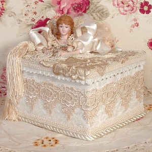May include: A white lace-trimmed box with a porcelain doll inside. The doll is wearing a white dress and has blonde hair. The box has a white tassel hanging from it.