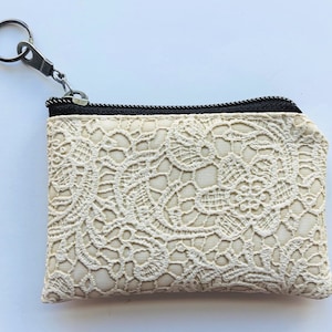 May include: A small, white lace zippered pouch with a silver key ring attached. The pouch has a black zipper and a delicate floral pattern.