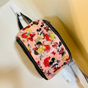 May include: A pink and black crossbody bag with a floral and Mickey Mouse print. The bag has a zipper closure and a long adjustable strap.