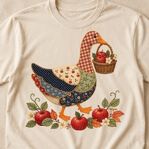 May include: A cream-colored t-shirt featuring a graphic of a goose with a patchwork-style body in red gingham, blue floral, and green leaf patterns. The goose carries a woven basket of red apples and stands among red apples and green leaves.