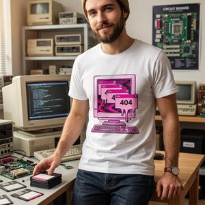 May include: A white t-shirt featuring a graphic print of a pink vintage computer monitor with a keyboard. The screen displays multiple overlapping error windows, including one clearly labeled with the text 404, with a melting effect design.