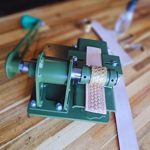 Könnte beinhalten: Eine grüne Lederprägemaschine aus Metall mit Handkurbel und einer zylindrischen Messingwalze mit Kreuzschraffur. Das Gerät ist dabei zu sehen, wie es eine dekorative Textur auf einen langen, hellbraunen Lederstreifen drückt.