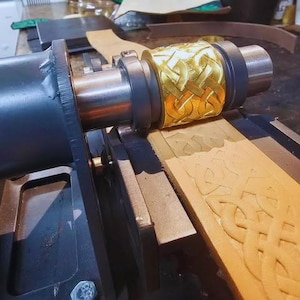 Könnte beinhalten: Eine Lederprägemaschine aus Metall mit einer goldfarbenen zylindrischen Walze, die ein komplexes keltisches Knotenmuster aufweist. Die Maschine drückt das geprägte Design in einen hellbraunen Lederstreifen und zeigt den Abdruck.