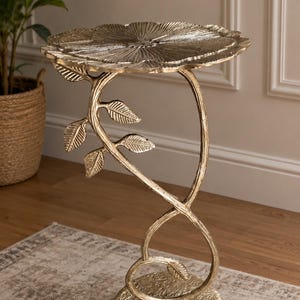 May include: A gold-colored metal side table featuring a circular top shaped like a flower with textured petals. The base is a solid circular disc, and the central support column is designed to resemble a vine with small leaves attached to the stem.