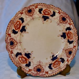 May include: A round ceramic plate with a scalloped edge, featuring a cream-coloured centre and a decorative border. The border displays a repeating pattern of orange floral motifs, dark blue accents, and intricate red scrollwork designs.