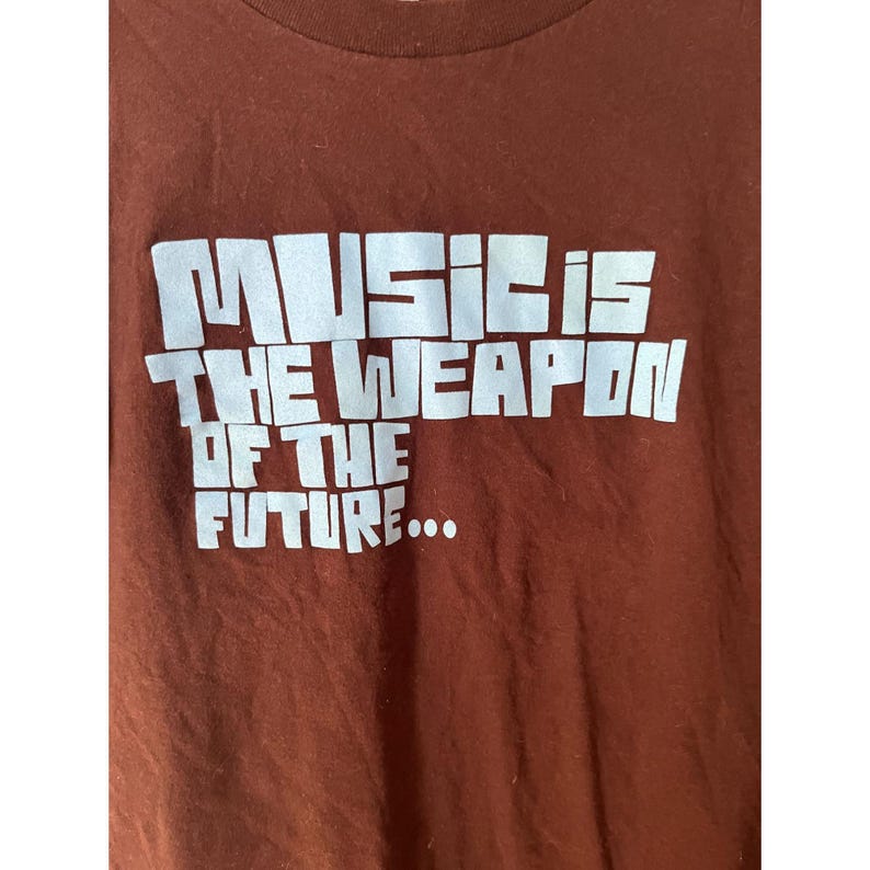 May include: A brown t-shirt with the text "MUSIC IS THE WEAPON OF THE FUTURE..." printed in white.