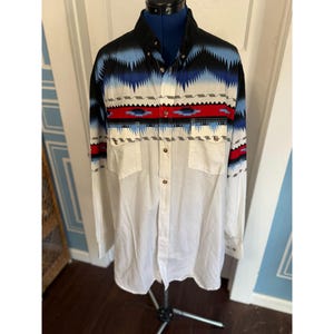 May include: A long-sleeved button-down shirt with a white body and a patterned yoke. The yoke features a geometric design in black, blue, red, and gray. The shirt has two chest pockets and a black collar.