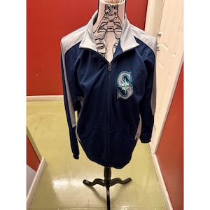 May include: A navy blue zip-up jacket with gray accents on the shoulders and collar. The jacket features the Seattle Mariners logo on the left chest. The jacket is displayed on a mannequin.