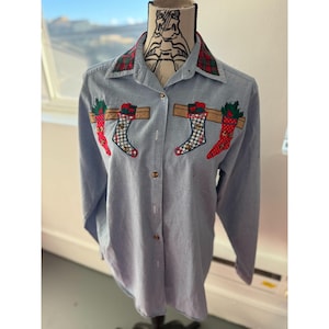 Könnte beinhalten: Hellblaues Button-Down-Hemd mit langen Ärmeln und einem karierten Kragen. Das Hemd hat gestickte Weihnachtsstrümpfe und Stechpalmen auf der Vorderseite. Die Strümpfe sind rot, grün und weiß und hängen an einem braunen Holzbalken.