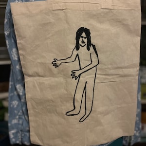 Puede incluir: Una bolsa de lona blanca con un diseño bordado en negro de una persona con el pelo largo y los brazos extendidos.