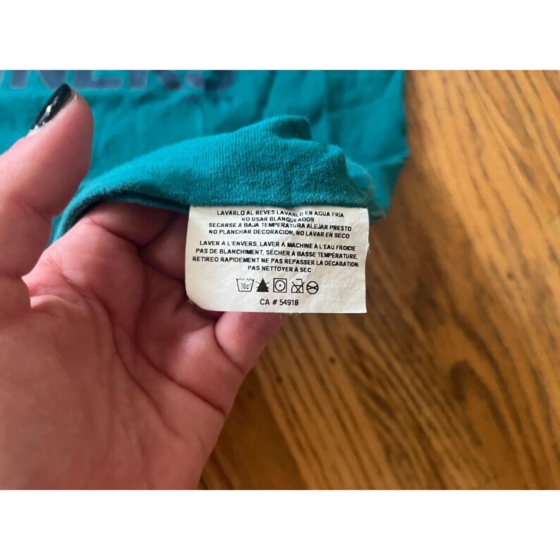 May include: A teal blue t-shirt with care instructions in Spanish and French. The instructions include washing, drying, and ironing recommendations. The tag also includes the text "CA # 54918".