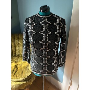 May include: A black sequined top with a geometric pattern in silver and pink. The top has long sleeves and a round neckline.