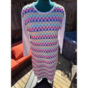 May include: A long-sleeved dress featuring a white base and a vibrant geometric pattern of triangles in red, blue, yellow, and green. The sleeves and neckline are white. The dress is displayed on a mannequin.