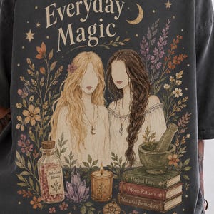May include: A charcoal gray t-shirt featuring a graphic print of two figures with long hair, surrounded by flowers, a bottle labeled Botanical Blends, a crystal, a candle, and a stack of books titled Herbal Lore, Moon Rituals, and Natural Remedies.