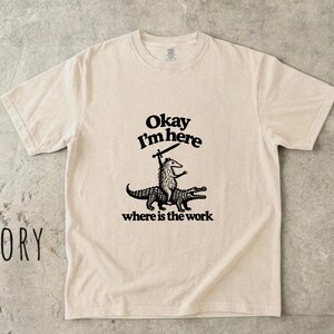 Puede incluir: Una camiseta de manga corta color marfil con un gráfico negro de un zarigüeya montando un caimán y sosteniendo una espada. El texto sobre el gráfico dice "Okay I'm here" y el texto debajo "where is the work". Se ve una etiqueta de 100% algodón.