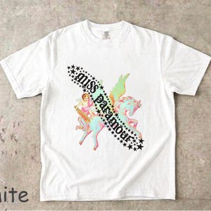 May include: A white short-sleeve t-shirt featuring a graphic print of a winged horse and a cherub. The design includes the text "miss paramour" written in a black gothic font, surrounded by a diagonal border of black stars. The shirt is 100% cotton.