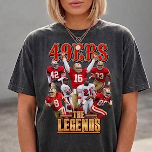 Könnte beinhalten: Ein anthrazitfarbenes Kurzarm-T-Shirt mit einem Grafikdruck von sechs Footballspielern in rot-goldenen Uniformen. Der Text 49ERS erscheint oben in Rot und THE LEGENDS ist unten im Grafikdesign in Gold aufgedruckt.