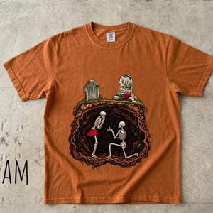 May include: A burnt orange short-sleeve t-shirt featuring a graphic of two skeletons inside an underground cavern beneath two tombstones. One skeleton wears a red skirt and the other kneels while holding a ring. The text 100% COTTON is in the corner.