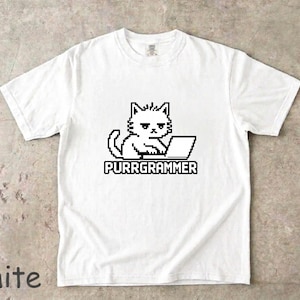 以下が含まれることがあります： ノートパソコンの前に座る猫の黒いピクセルアートグラフィックが描かれた白い半袖Tシャツ。猫の下には、太いブロック状のピクセルフォントでPURRGRAMMERという文字がプリントされています。隅にある小さな黒いラベルには100% COTTONと記載されています。