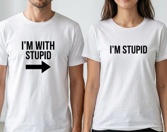 Puede incluir: Dos camisetas blancas de manga corta. La camiseta de la izquierda tiene el texto negro I'M WITH STUPID y una flecha negra apuntando a la derecha. La camiseta de la derecha tiene el texto negro I'M STUPID. Ambas tienen cuello redondo.