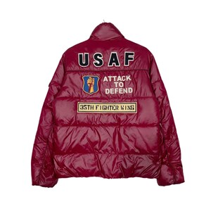 May include: A shiny maroon puffer jacket featuring back patches with the text USAF, a shield emblem with a fist holding a dagger, the text ATTACK TO DEFEND, and a rectangular patch reading 35TH FIGHTER WING. The jacket has a high collar and quilted design.