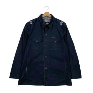Könnte beinhalten: Eine dunkelblaue Jacke mit Knopfleiste, klassischem Kragen, zwei Brusttaschen mit Knopfklappen und Schulterklappen mit weißen Streifendetails. Das Innenfutter des Kragens zeigt ein beiges, schwarzes und weißes Karomuster.