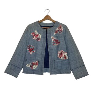 May include: A light blue, textured jacket with a collarless, open front design. The garment features embroidered floral patches in red, white, and black thread. It includes two front patch pockets, long sleeves, and vertical stitching details.
