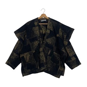 Puede incluir: Una chaqueta abierta con un patrón de patchwork texturizado en azul marino oscuro y bronce dorado. Esta prenda tiene mangas anchas, cuello en V y dos bolsillos delanteros. La chaqueta se muestra en una percha de madera sobre fondo blanco.