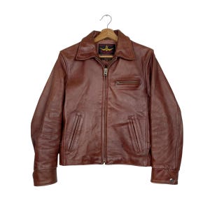 May include: A brown leather jacket featuring a pointed collar, a full-length front zipper, and two vertical zippered pockets at the waist. A single horizontal zippered pocket is located on the upper left chest. The interior label reads Aero Leather.