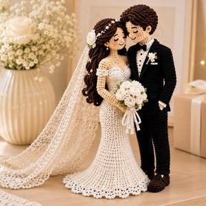 Pode incluir: Um par de bonecos de crochê representando um casal de noivos. A figura à esquerda veste um vestido branco com ombros à mostra, um longo véu de renda e segura um buquê de flores brancas. A figura à direita veste um terno preto com camisa.