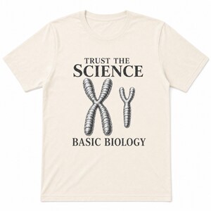 May include: A cream-colored short-sleeve t-shirt featuring a black graphic print. The design includes the text "TRUST THE SCIENCE" above a drawing of X and Y chromosomes, with the text "BASIC BIOLOGY" printed below the chromosome illustration.