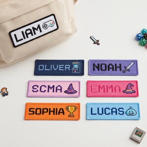 May include: A beige canvas backpack with a rectangular patch reading LIAM and a game controller icon. Six additional rectangular patches feature names and pixel art icons: OLIVER, NOAH, SCMA, EMMA, SOPHIA, and LUCAS, alongside small game-themed pins.