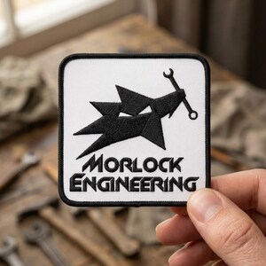 May include: A square embroidered patch with a white background and a black border. The design features a black geometric shape resembling a stylized animal head holding a wrench, with the text MORLOCK ENGINEERING printed in black below.
